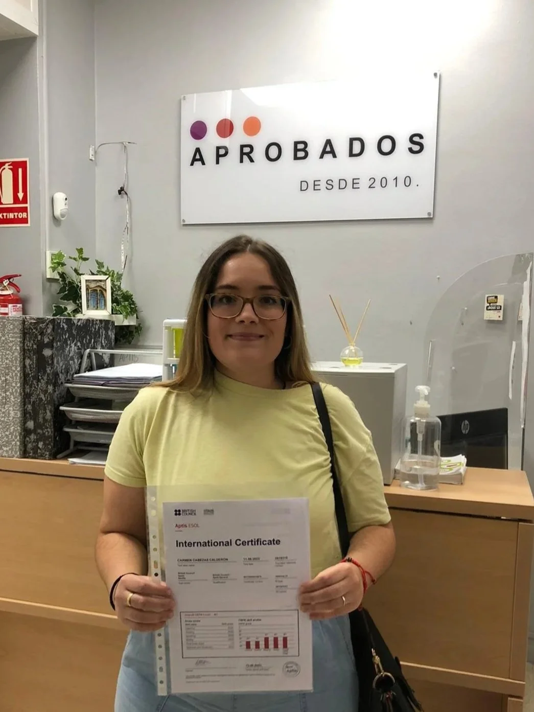 Mujer sonriendo frente a un mostrador en una oficina, sosteniendo un certificado internacional y en el fondo un cartel que dice 'Aprobados desde 2010'.