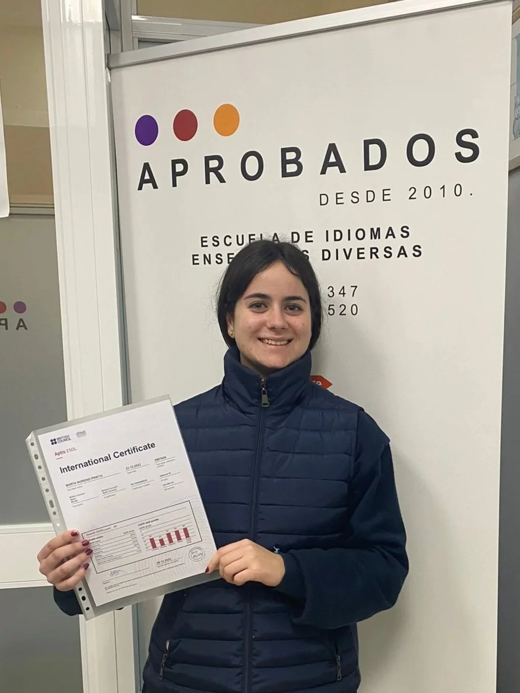 Mujer sonriendo sosteniendo un certificado internacional en un aula de idiomas, frente a un cartel que dice "Aprobados desde 2010" y "Escuela de idiomas enseñanza diversa".