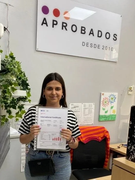Joven mujer sosteniendo un certificado internacional en una oficina, con cartel que dice 'Aprobados desde 2010' en la pared.