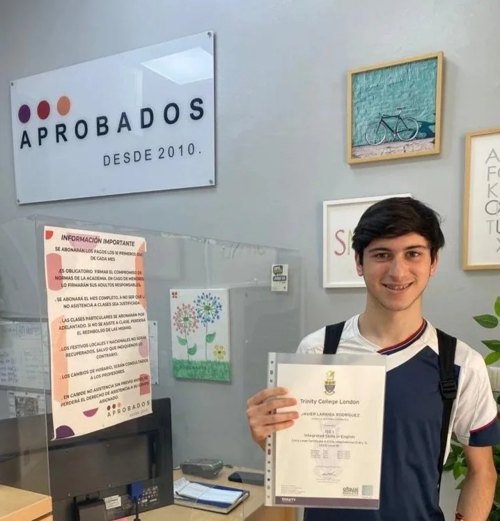 Joven sonriendo en recepción de oficina, sosteniendo un documento y con cartel y cuadros decorativos en la pared.