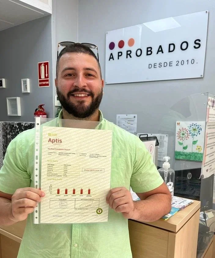 Hombre sonriendo sosteniendo un certificado en una oficina, con un cartel que dice 'APROBADOS DESDE 2010' al fondo.