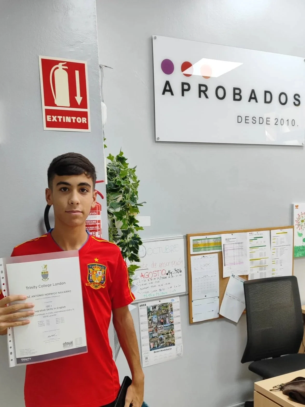 Joven sosteniendo un certificado en una oficina con carteles y notas en la pared.