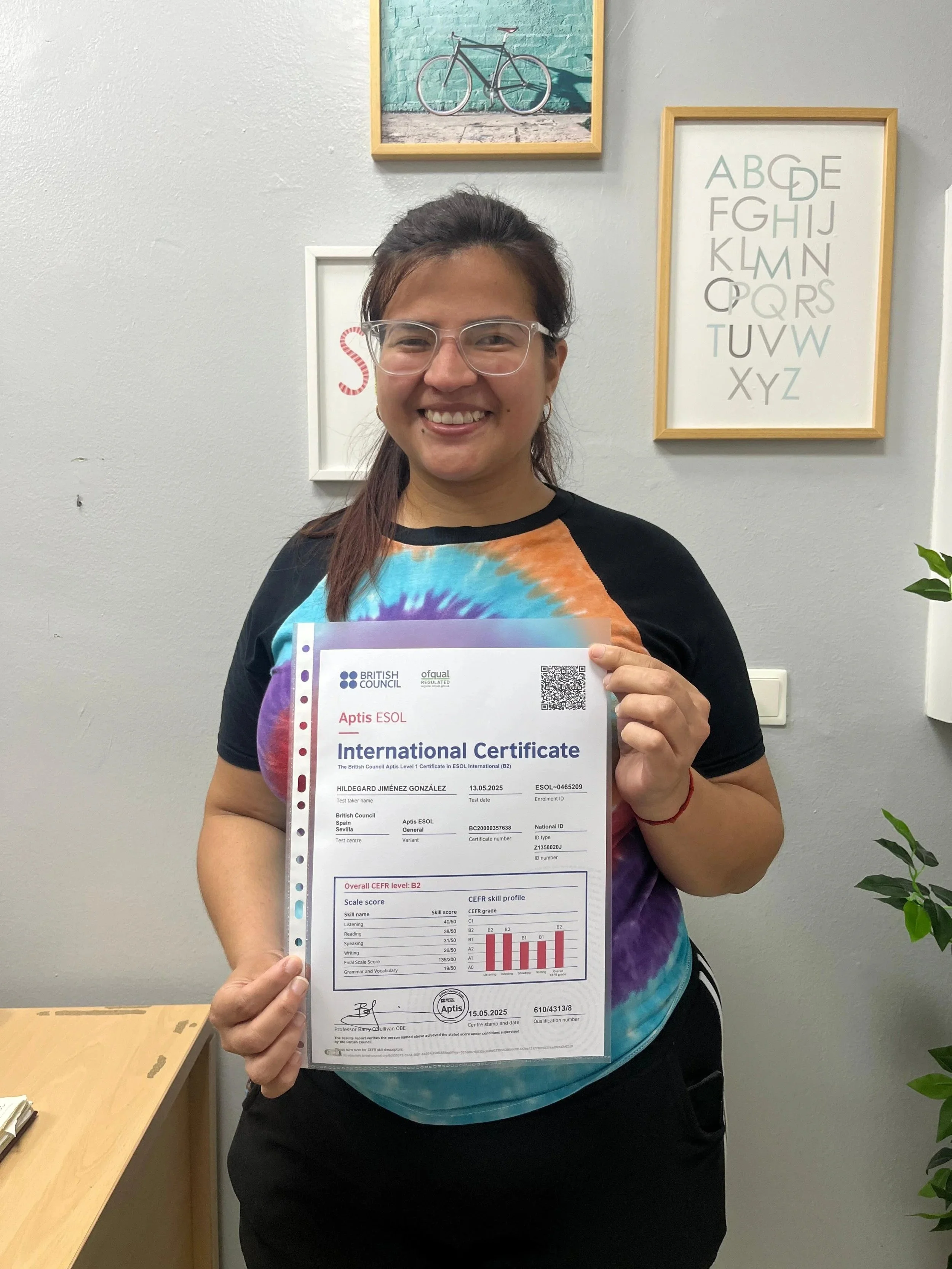 Mujer sonriendo sosteniendo un certificado de idioma en un ambiente con cuadros en la pared.