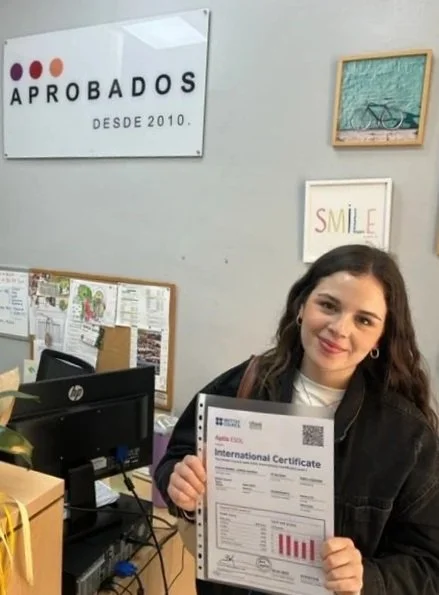 Mujer sonriendo sosteniendo un certificado internacional en una oficina. En la pared, carteles que dicen 'APROBADOS' y 'SMILE'.