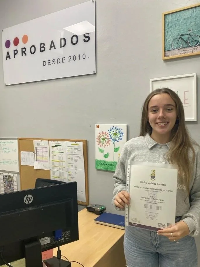 Una joven sonriendo sosteniendo un certificado en una oficina, con un cartel que dice 'APROBADOS' y decoraciones en la pared.