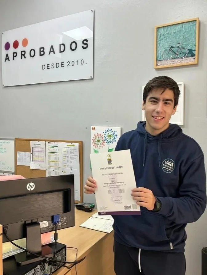 Estudiante sosteniendo certificado en departamento de oficina con cartel que dice 'Aprobados desde 2010' en la pared.