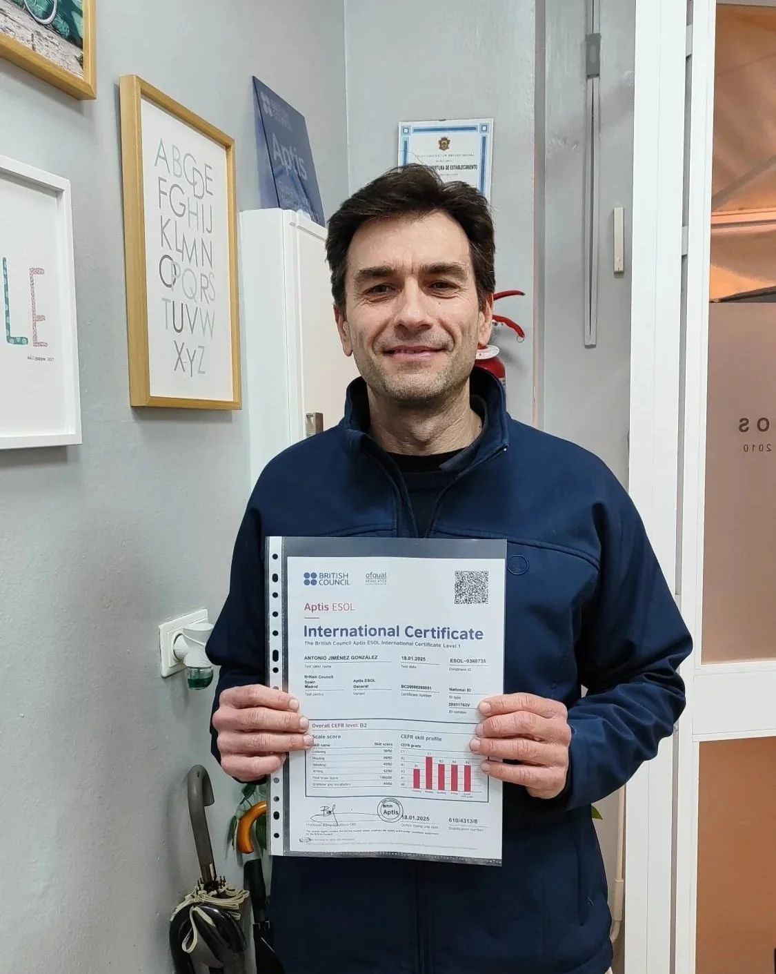 Hombre sonriendo, sosteniendo un certificado internacional en una oficina, con paredes decoradas con obras de arte y certificados.