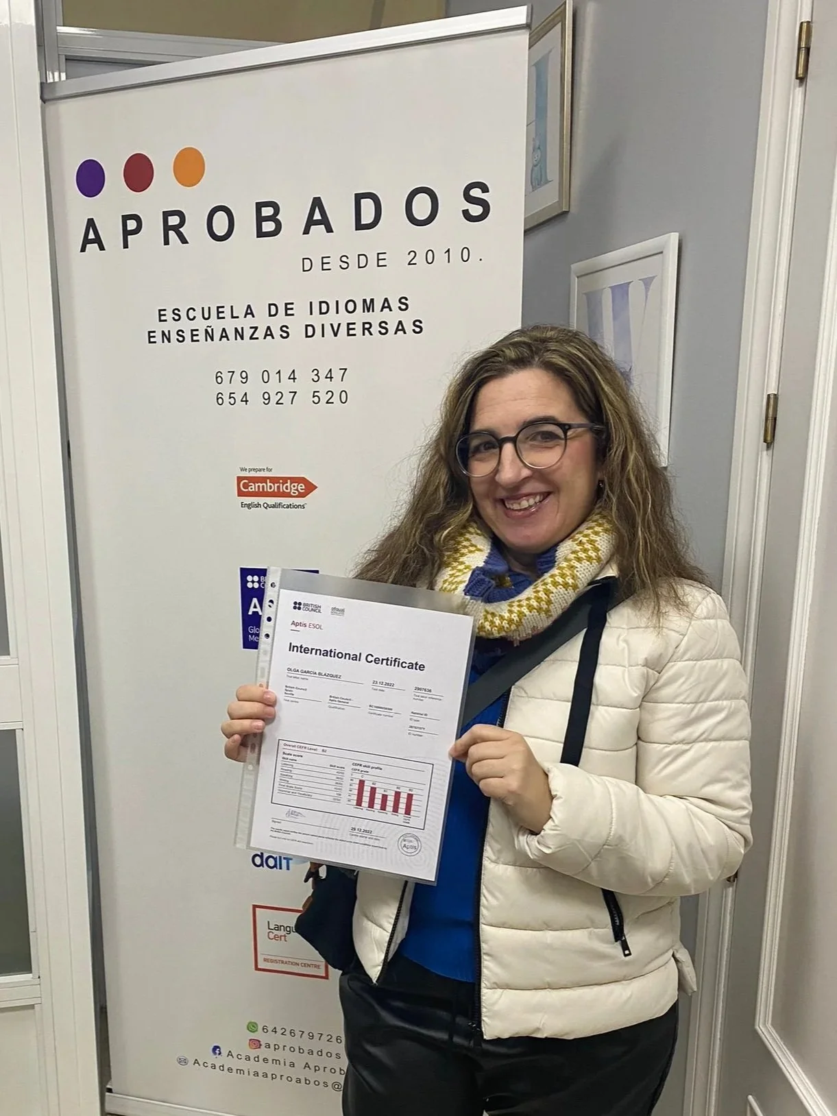 Mujer sonriendo, sosteniendo certificado internacional, de pie frente a cartel de escuela de idiomas y enseñanzas diversas.