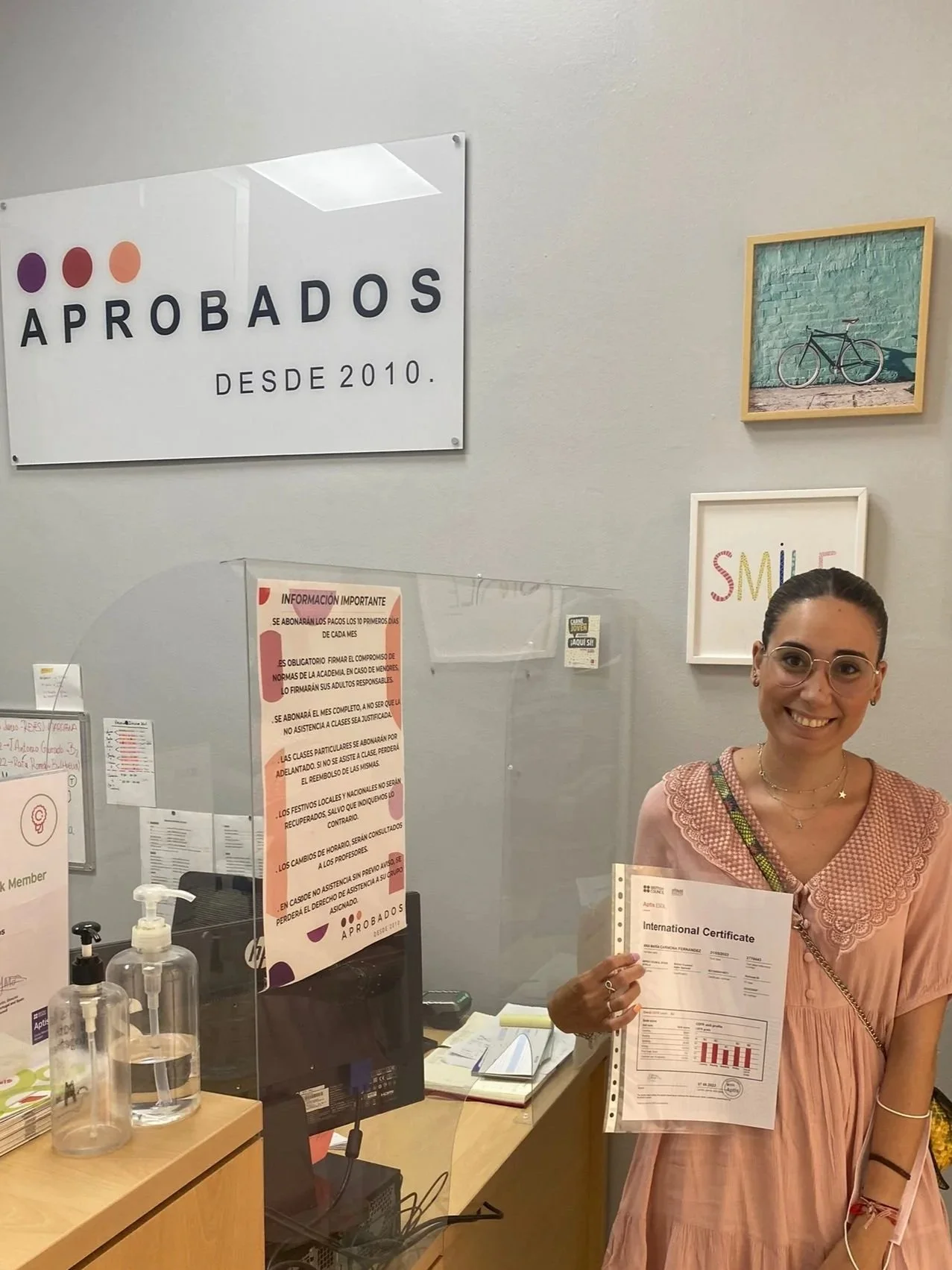 Mujer sonriendo en oficina, sosteniendo certificado internacional, detrás de escritorio con envases de gel y documentos, cartel que dice 'aprobados desde 2010' en la pared.