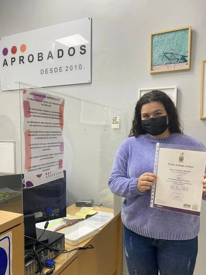 Joven sonriente con mascarilla negra, sosteniendo un diploma de la Trinity College London en un aula, con cartel y cuadros en la pared.