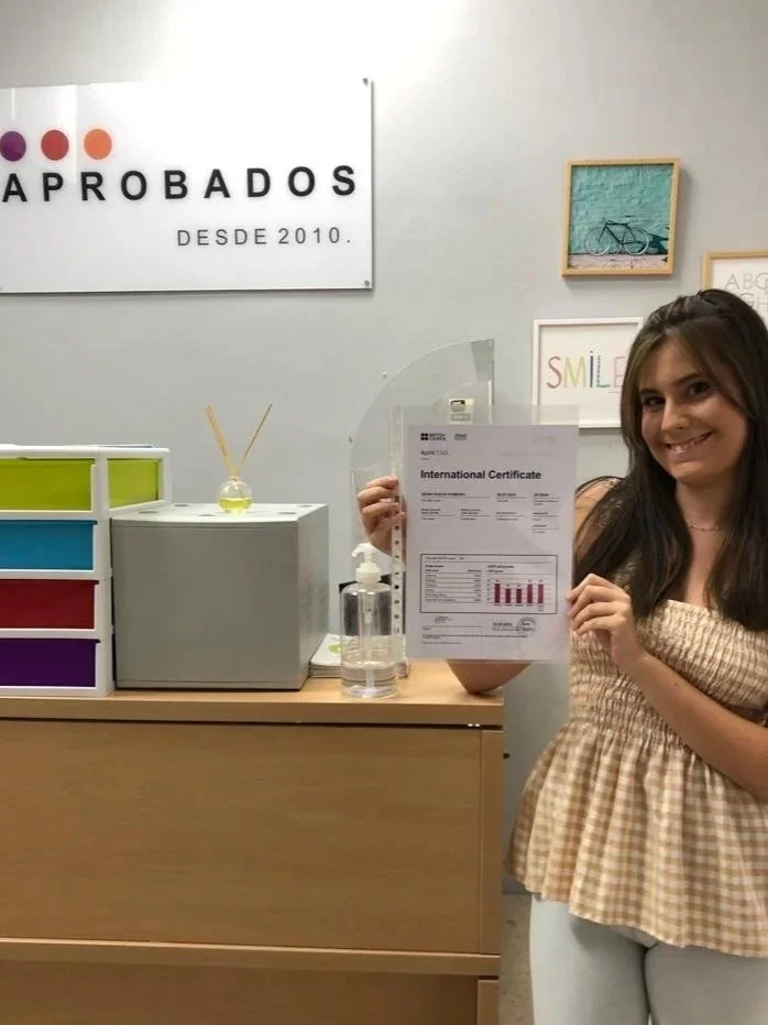 Una joven sosteniendo un certificado internacional en una oficina, con un cartel que dice 'APROBADOS' y decoraciones en la pared.