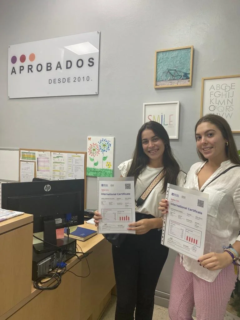 Dos jóvenes sosteniendo certificados en una oficina, con cartel que dice 'APROBADOS DESDE 2010', cuadros decorativos en la pared, y escritorio con ordenador y documentos.