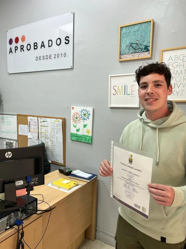 Joven sonriente sosteniendo un certificado en la academia, pared decorada con cuadros y cartel que dice 'APROBADOS DESDE 2010'.