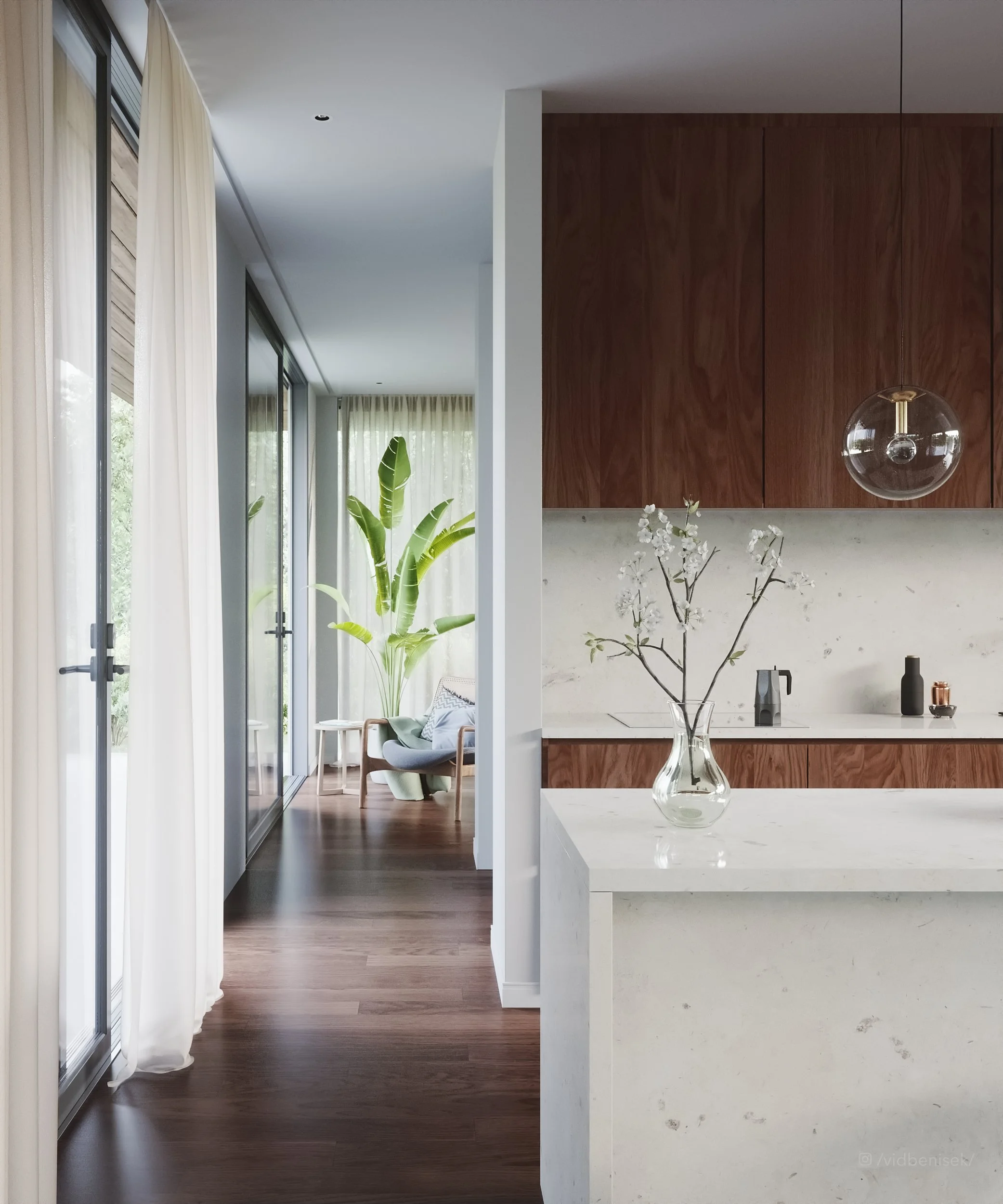 Interior view of a modern home with a white kitchen counter, a vase with branches, wooden cabinets, a hanging glass light, green houseplant, and a sitting area with a chair and curtains in the background.