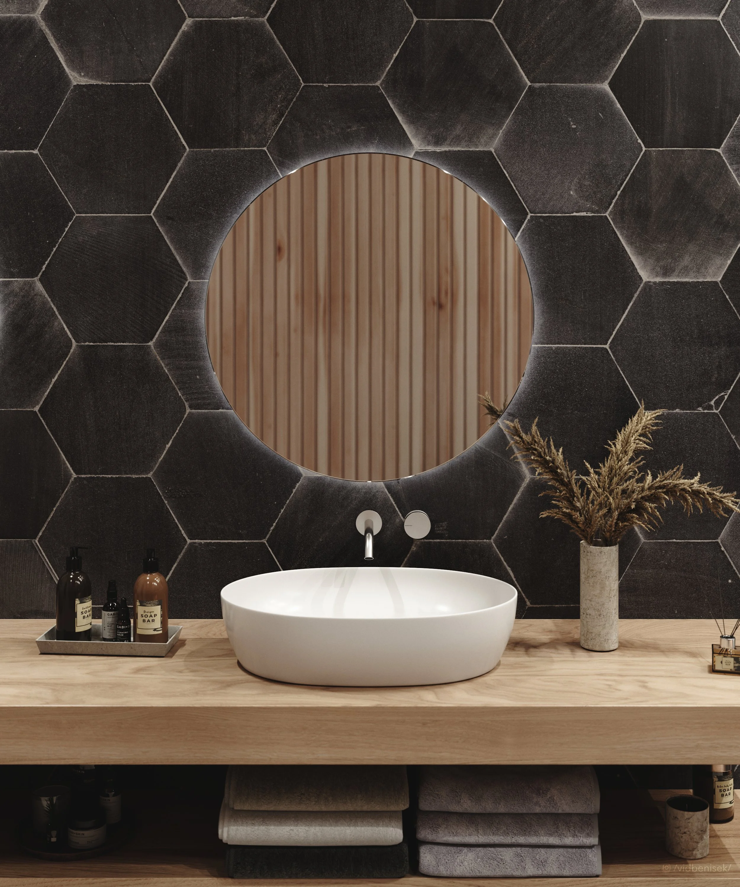 Modern bathroom vanity with a round mirror, white vessel sink, and dark tile hexagon wall, with bottles of soap and dried plants on wooden countertop.