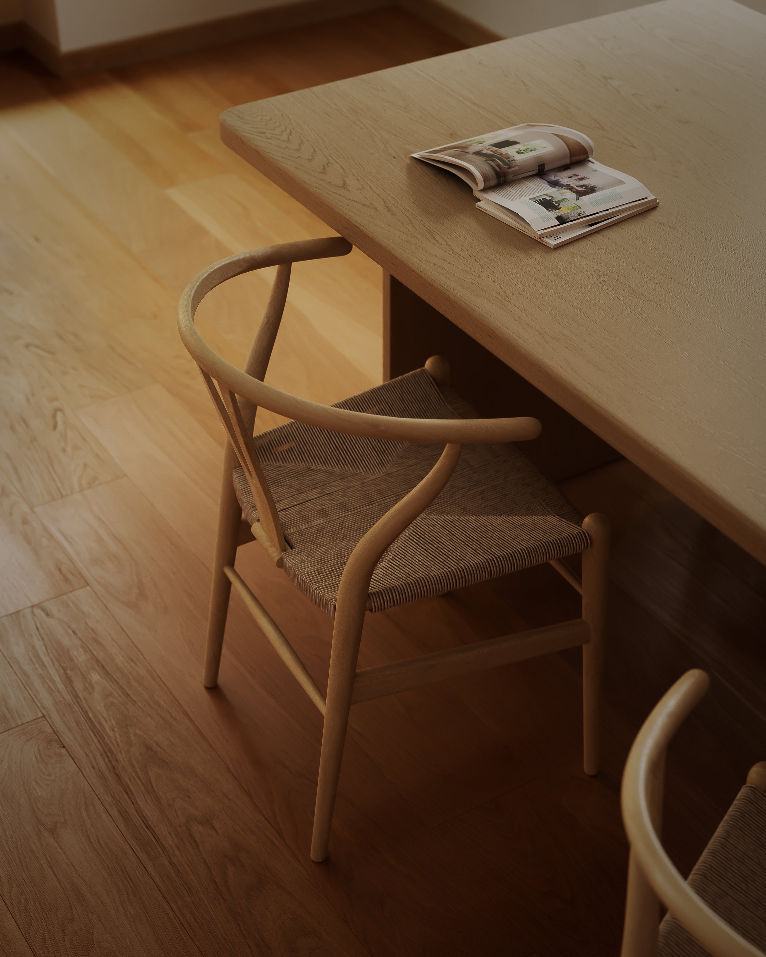 A wooden chair with a woven seat pulled out from a wooden table, with an open magazine on the table.