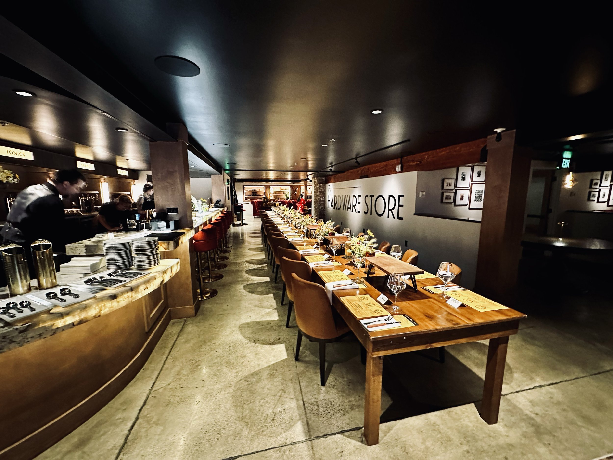 a private room for a gathering with ambient lighting, a bar with underlit countertop, a long table with brown leather chairs, set with glassware, menus, and flowers