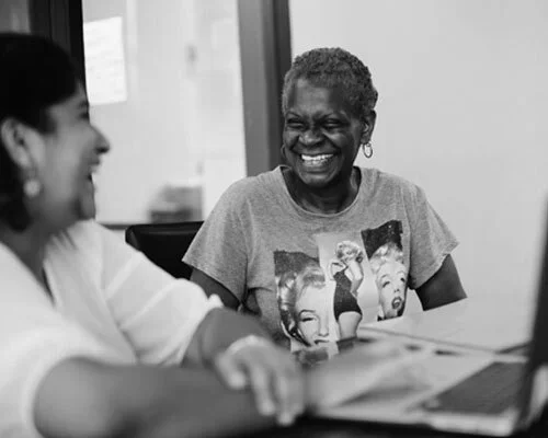 Two women laughing and talking at a table in an office setting.