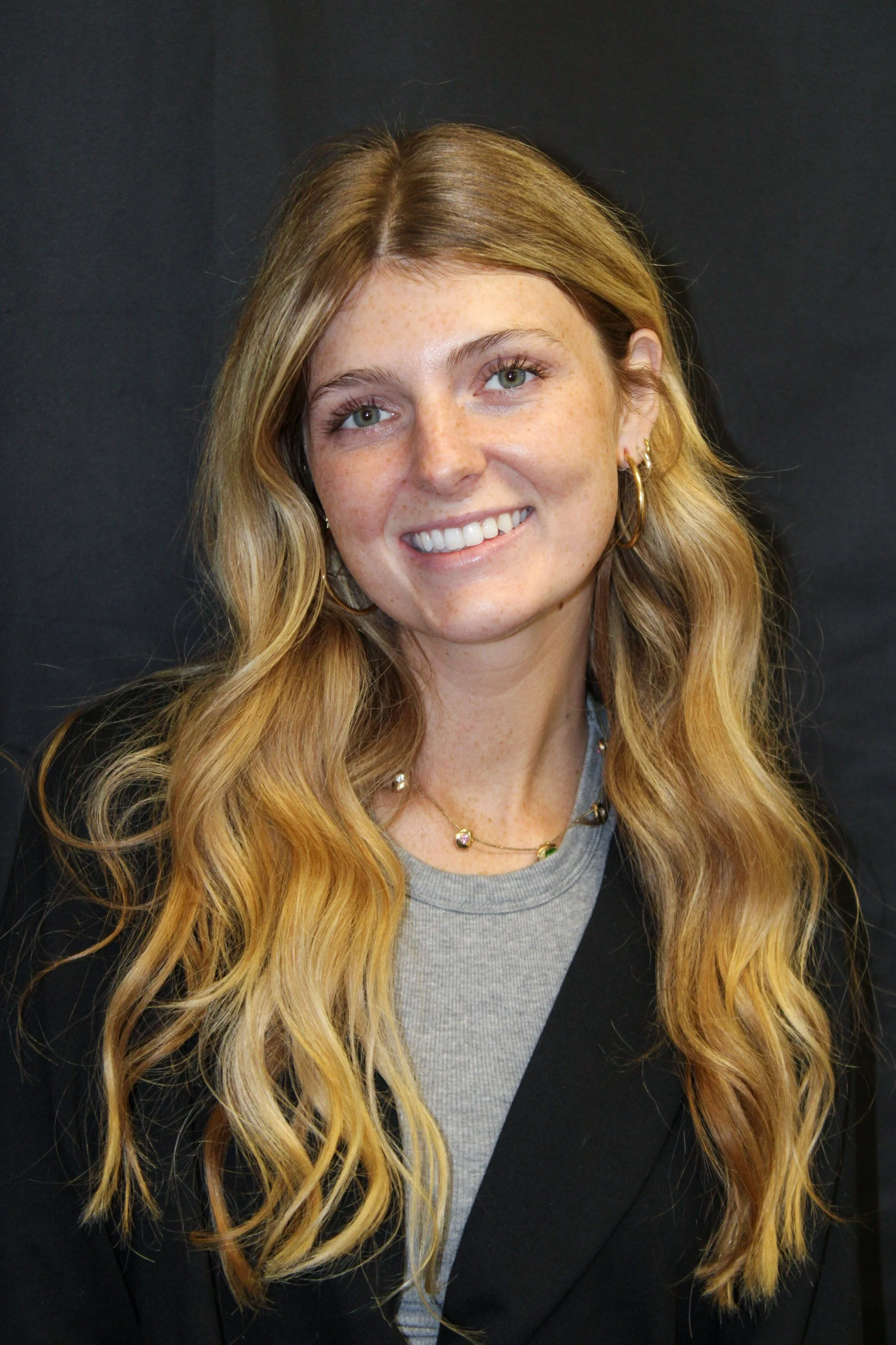 Portrait of a young woman with long, wavy blonde hair, smiling, wearing a black blazer, gray shirt, and gold jewelry, standing against a dark background.