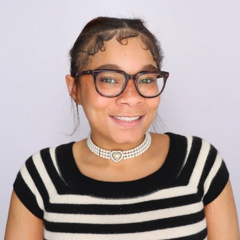 Young woman with dark hair in braids, wearing glasses, a pearl choker necklace, and a black and white striped shirt, smiling at the camera against a plain background.