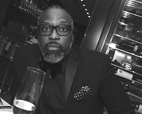 A man with glasses and a beard wearing a suit and bowtie, sitting at a table in a restaurant or bar, with a glass of champagne in front of him, in a black and white photo.