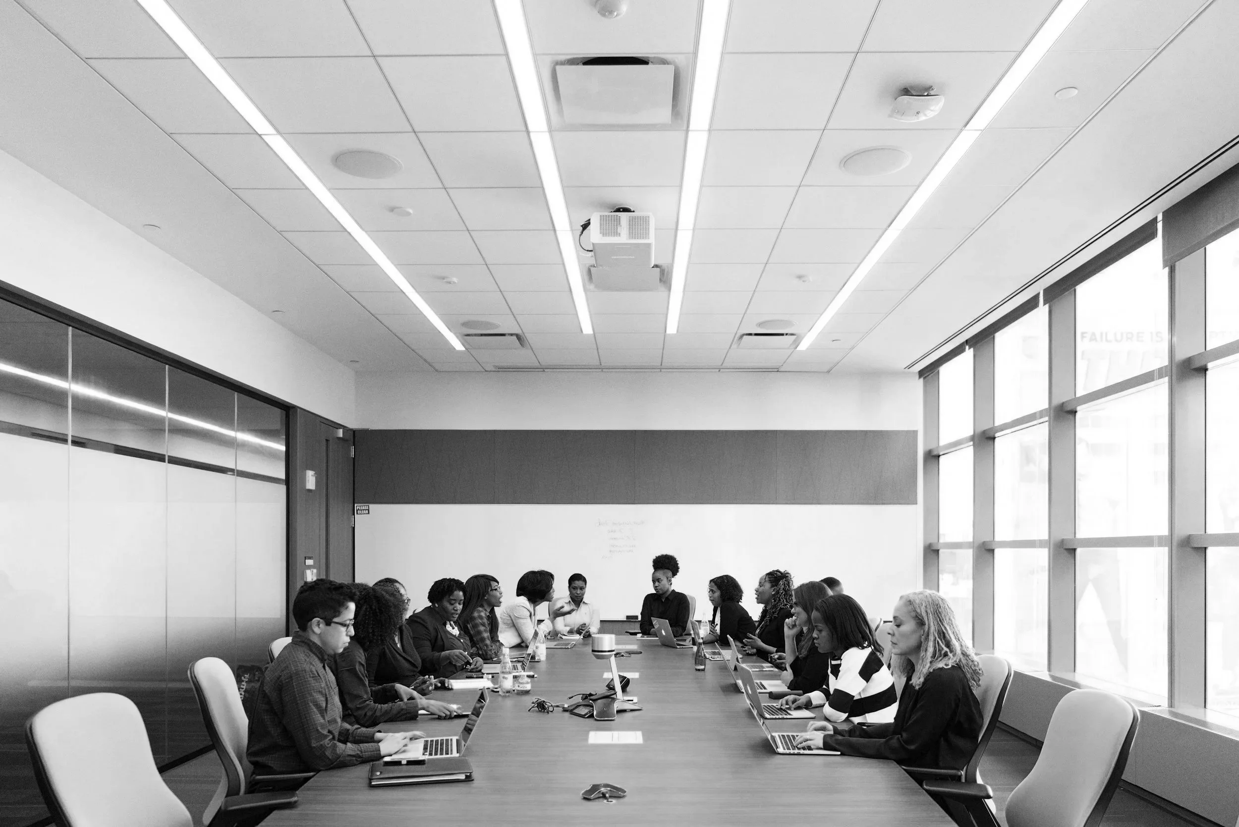 A diverse group of professionals in a modern conference room engaged in a meeting, with laptops open in front of them, during the daytime.