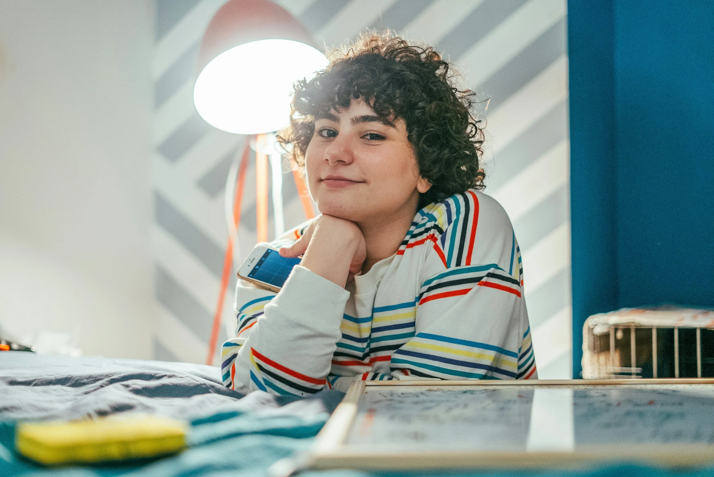 A young person with curly hair and a septum piercing, wearing a striped shirt, smiling and resting their chin on their hand while sitting at a desk, with a smartphone in hand, in a brightly lit room with a patterned wall and a lamp in the background.