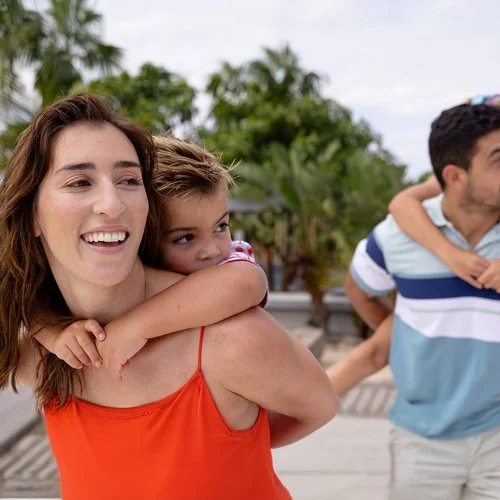 A woman smiling as she carries a young girl on her back, with another man partially visible in the background at an outdoor location with trees.