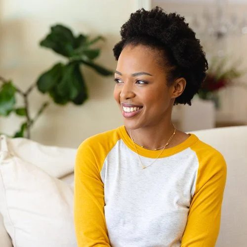 A smiling woman with short curly hair, wearing a yellow and white long-sleeve shirt, sitting on a sofa in a bright room with plants in the background.