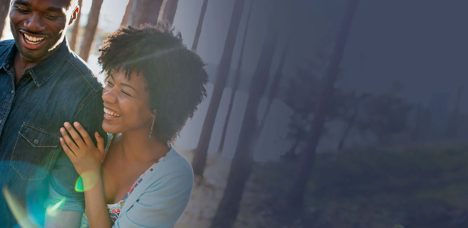 A smiling man and woman outdoors in a wooded area, enjoying each other's company during daylight.