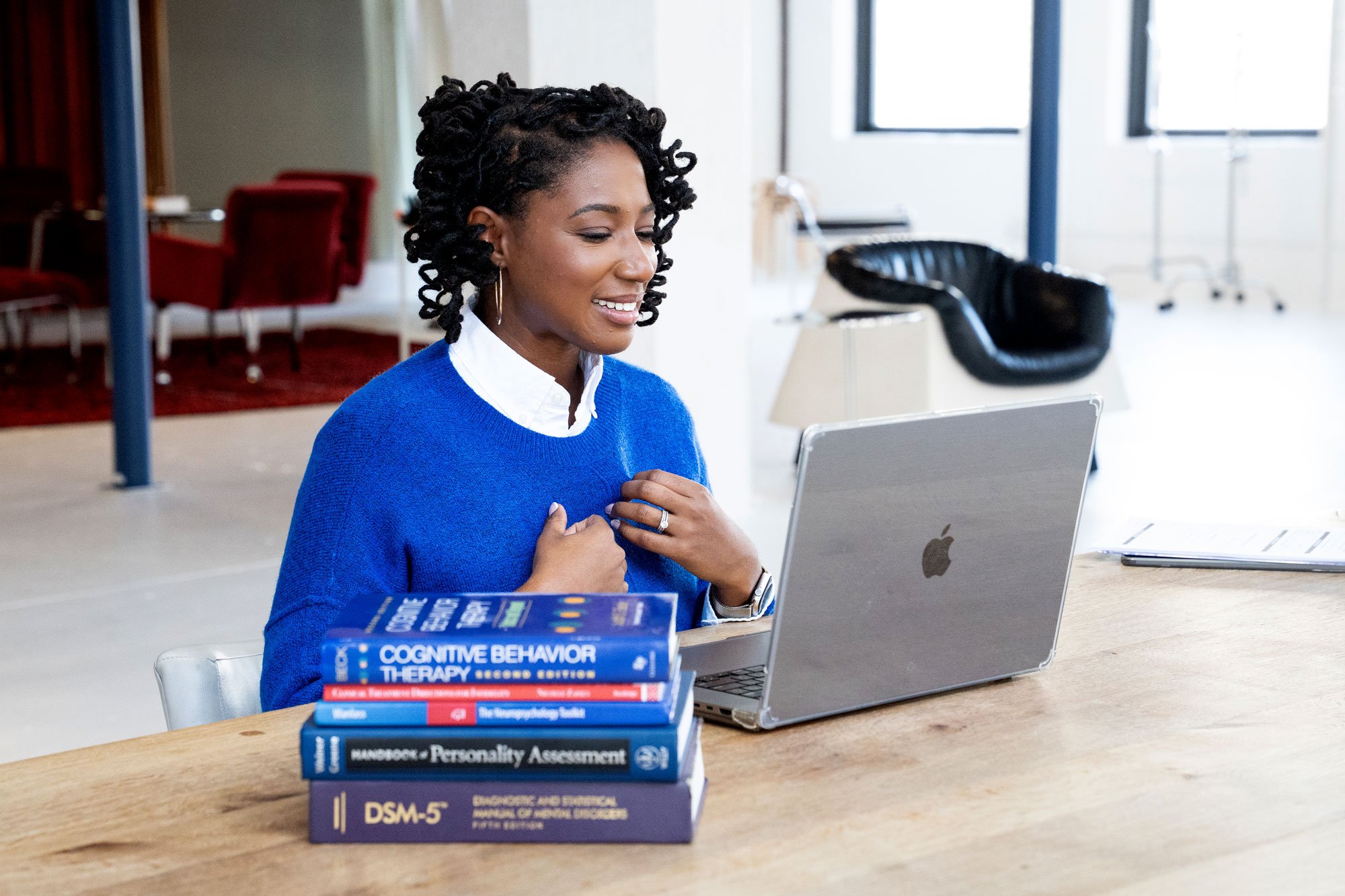 A woman sitting at a wooden table with a laptop in front of her, smiling and touching her chest. There are several books on mental health and psychology stacked on the table, including titles like 'Cognitive Behavior Therapy' and 'DSM-5'. The background features a modern office setting with windows and chairs.