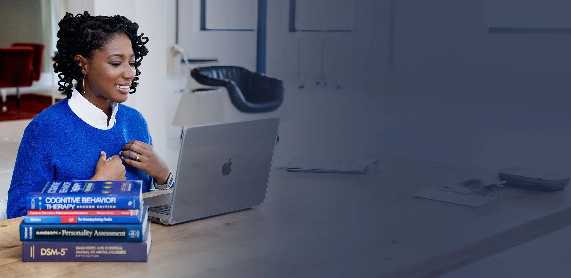 A woman sitting at a desk, smiling and looking at a laptop. A stack of books related to psychology, personality, and behavior assessment is on the desk. The background shows an office setting with a phone, papers, and a lamp.