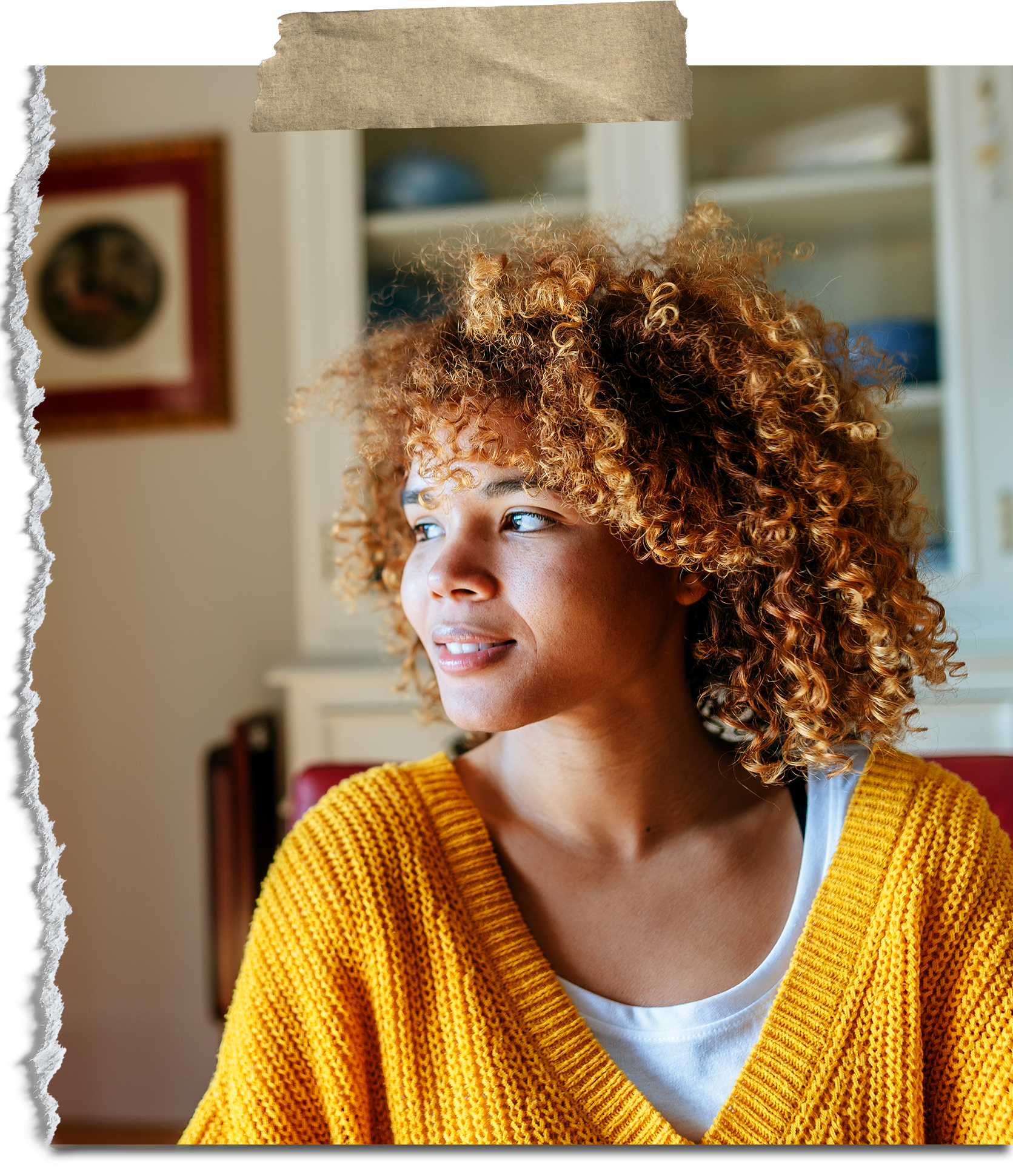 A woman with curly red hair sitting indoors, wearing a yellow knitted sweater and looking to her left.