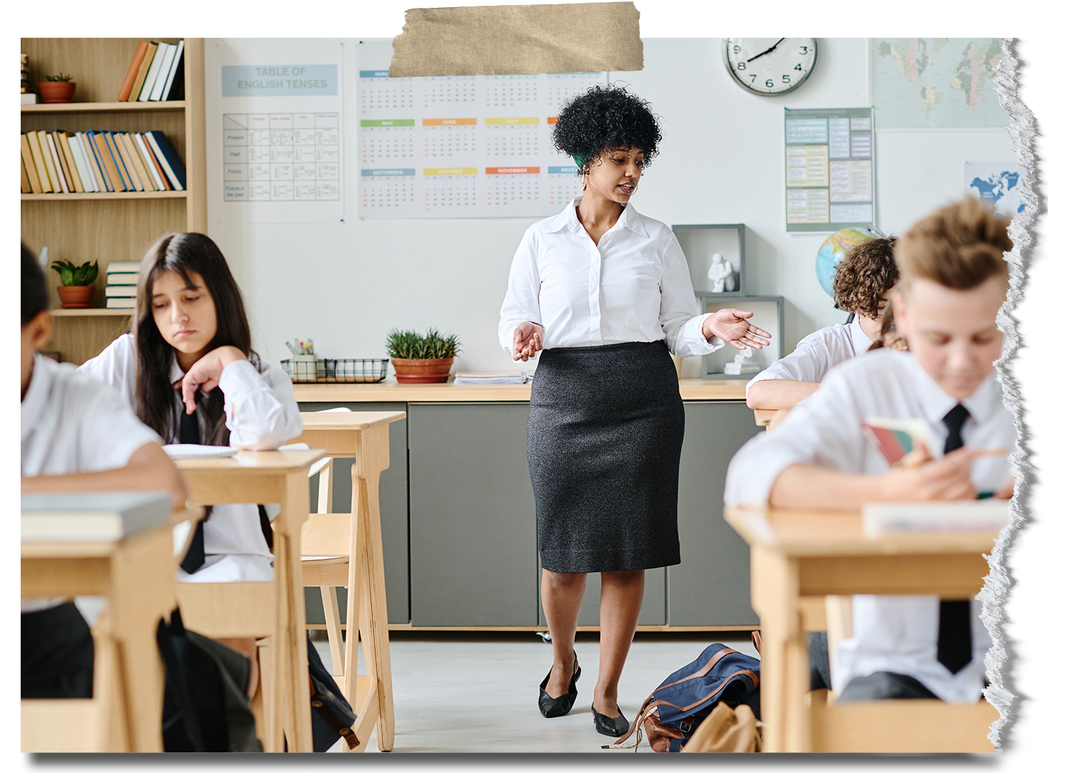Teacher standing in front of a classroom with students, some taking notes and others reading, in a classroom with maps and charts on the wall.