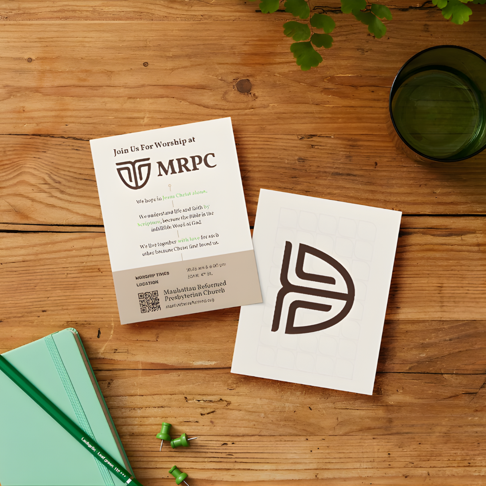 Flyer for MRPC church with a logo, a QR code, a white paper with a logo, a green notebook, green push pins, a green glass of water, and a leafy plant on a wooden table.