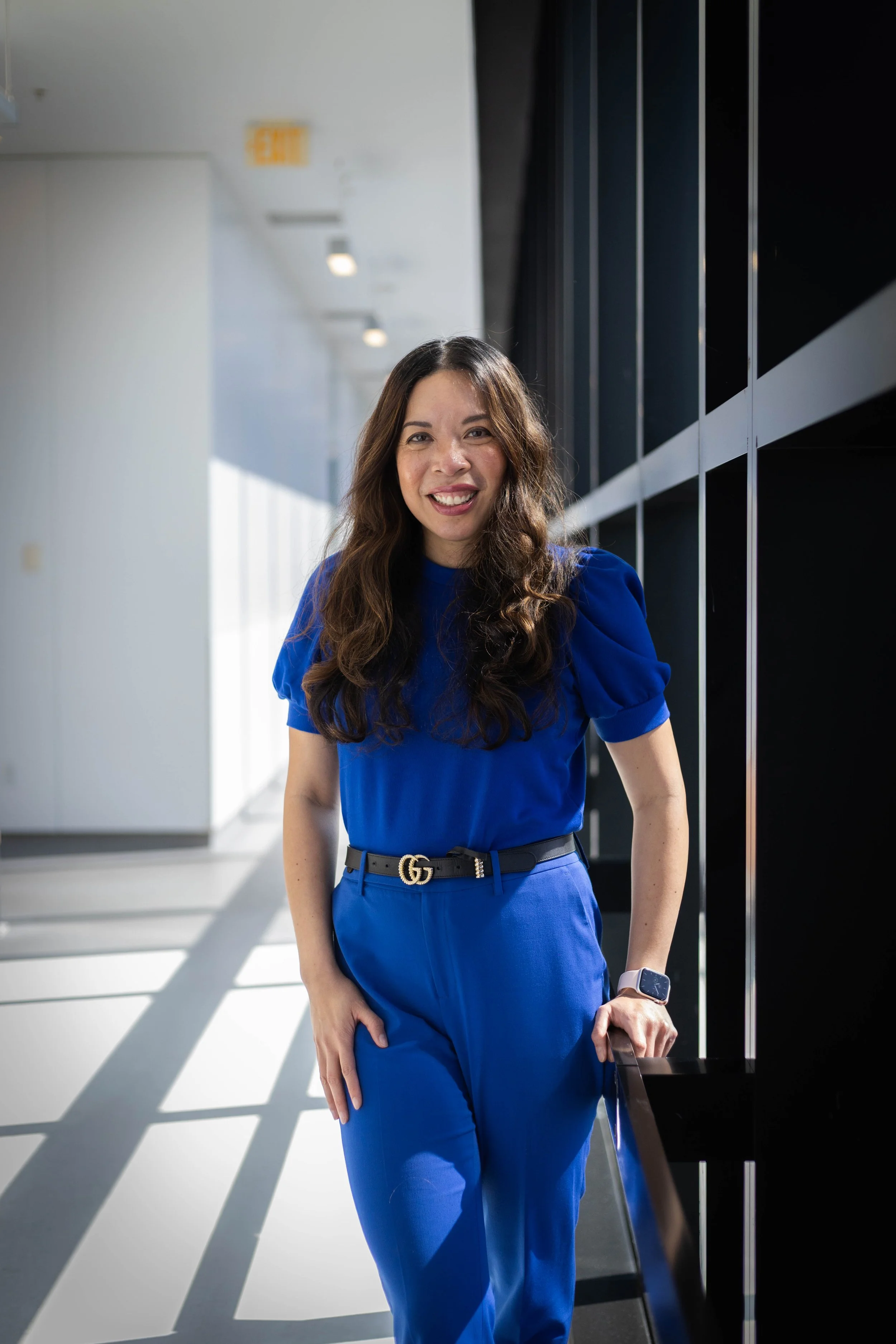 A woman with long wavy brown hair wearing a royal blue dress and a Gucci belt, standing in a modern hallway with black wall panels and windows, smiling at the camera, with a smartwatch on her left wrist.