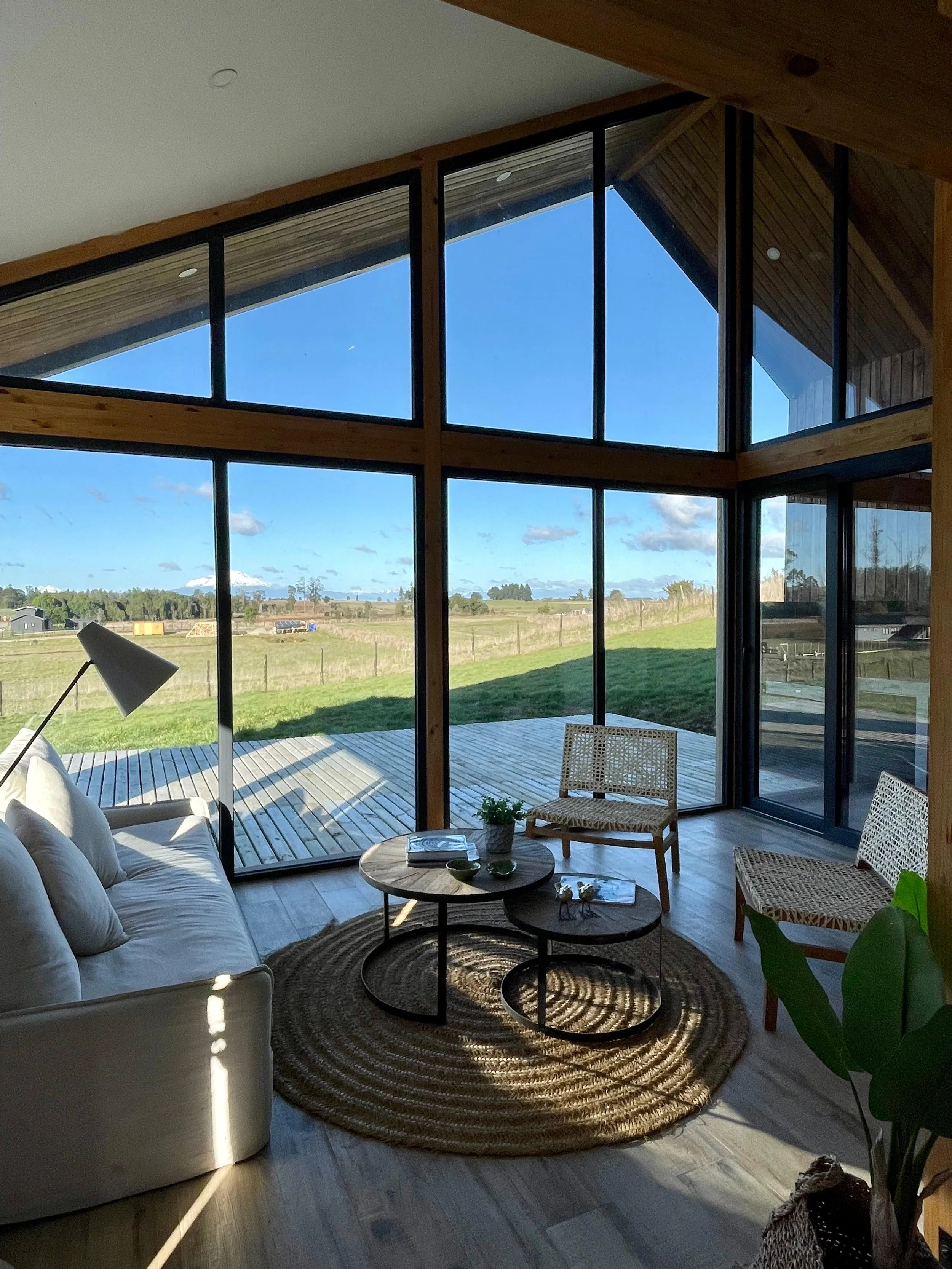 Vista de una sala moderna con grandes ventanales que muestran un paisaje campestre, con sillón blanco, sillas de mimbre, mesa de centro de madera y alfombra redonda de fibras naturales.