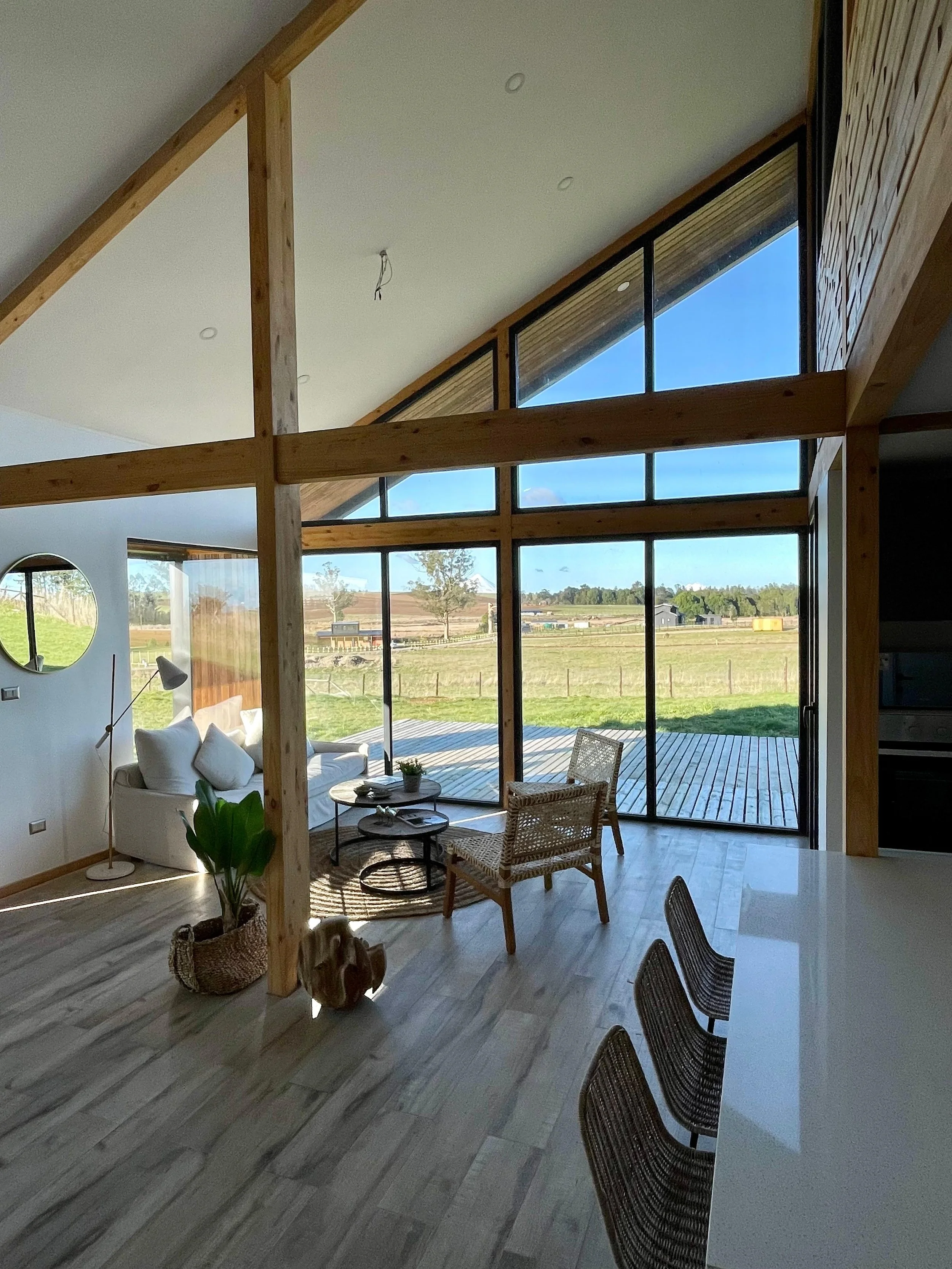 Interior de una casa moderna con paredes de vidrio grandes, con vista a un campo abierto. Tiene una sala de estar con sillones blancos, una planta en una cesta, una lámpara de lectura y una mesa de centro. Una zona de comedor con sillas de mimbre y u