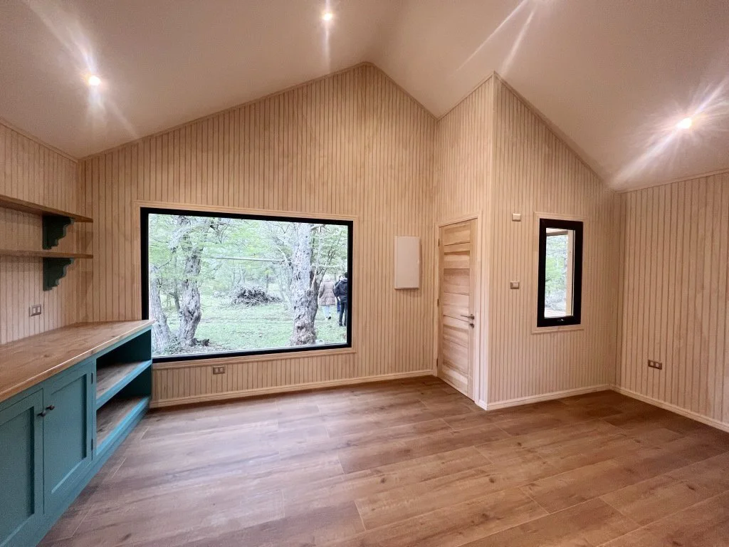 Sala con paredes de madera clara, ventanas grandes que muestran árboles y un espacio exterior natural, techo inclinado y pisos de madera