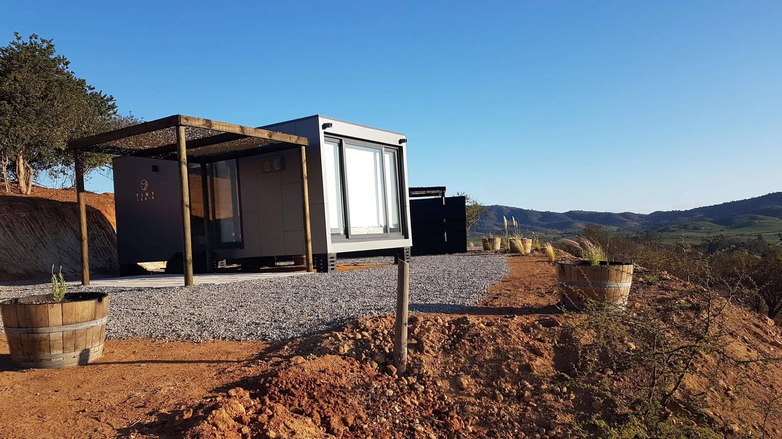 Tiny cabin en un paisaje rural con monte y cielo despejado, rodeada de plantas en jarrones de madera.