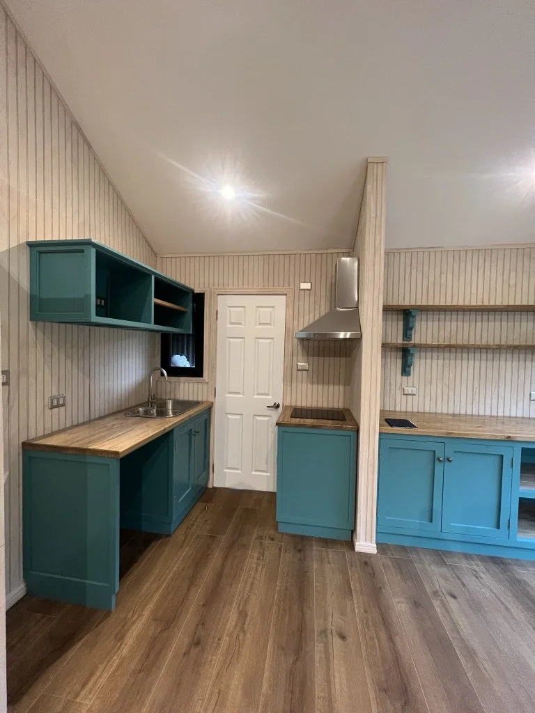 Cocina pequeña con gabinetes azules, encimera de madera, fregadero, estanterías abiertas, y una campana extractora, en una habitación con paredes de madera clara y suelo de madera.