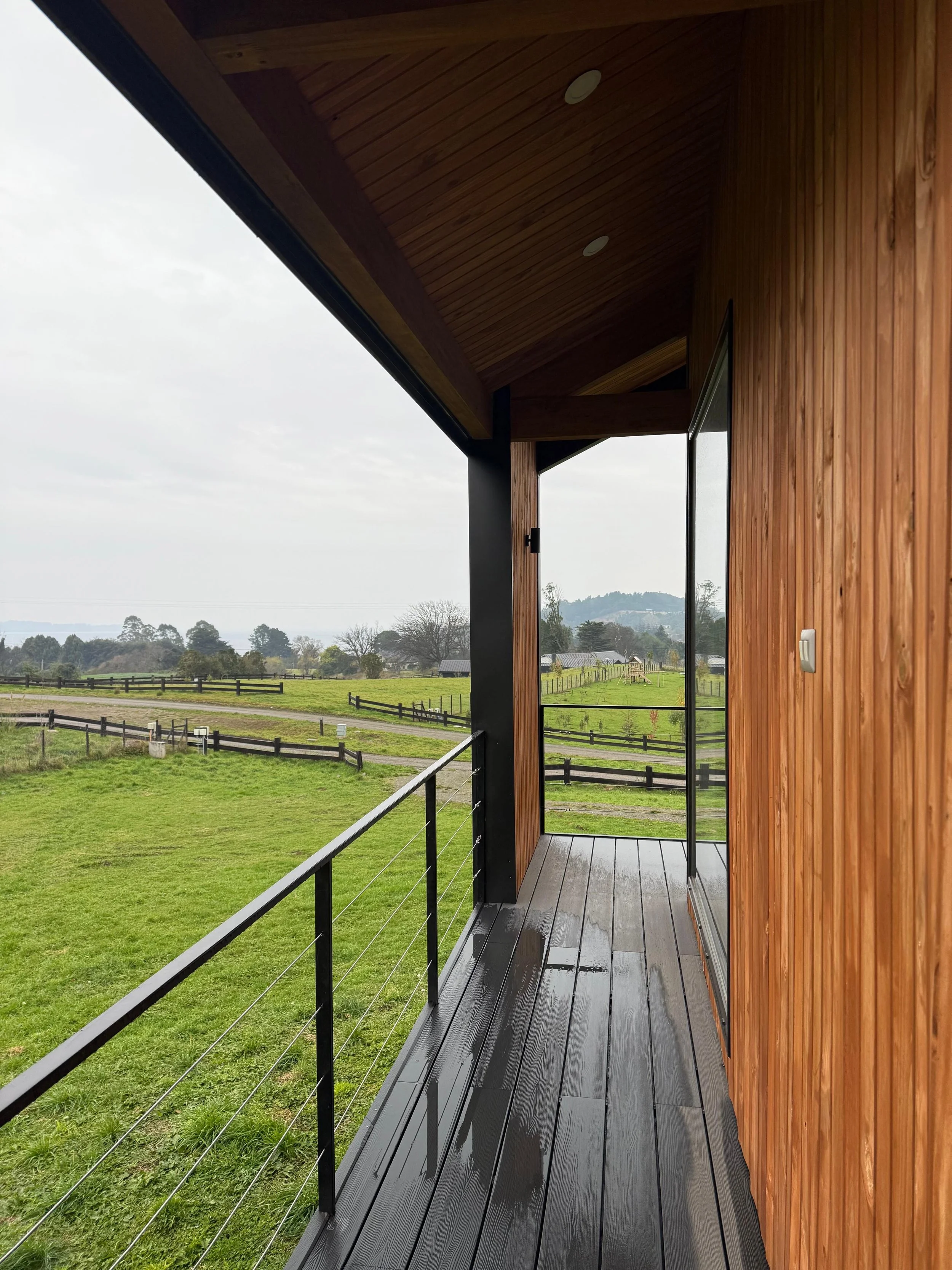 Vista de un balcón de madera con barandal de cables en un entorno rural con praderas verdes y árboles, con cielo nublado.