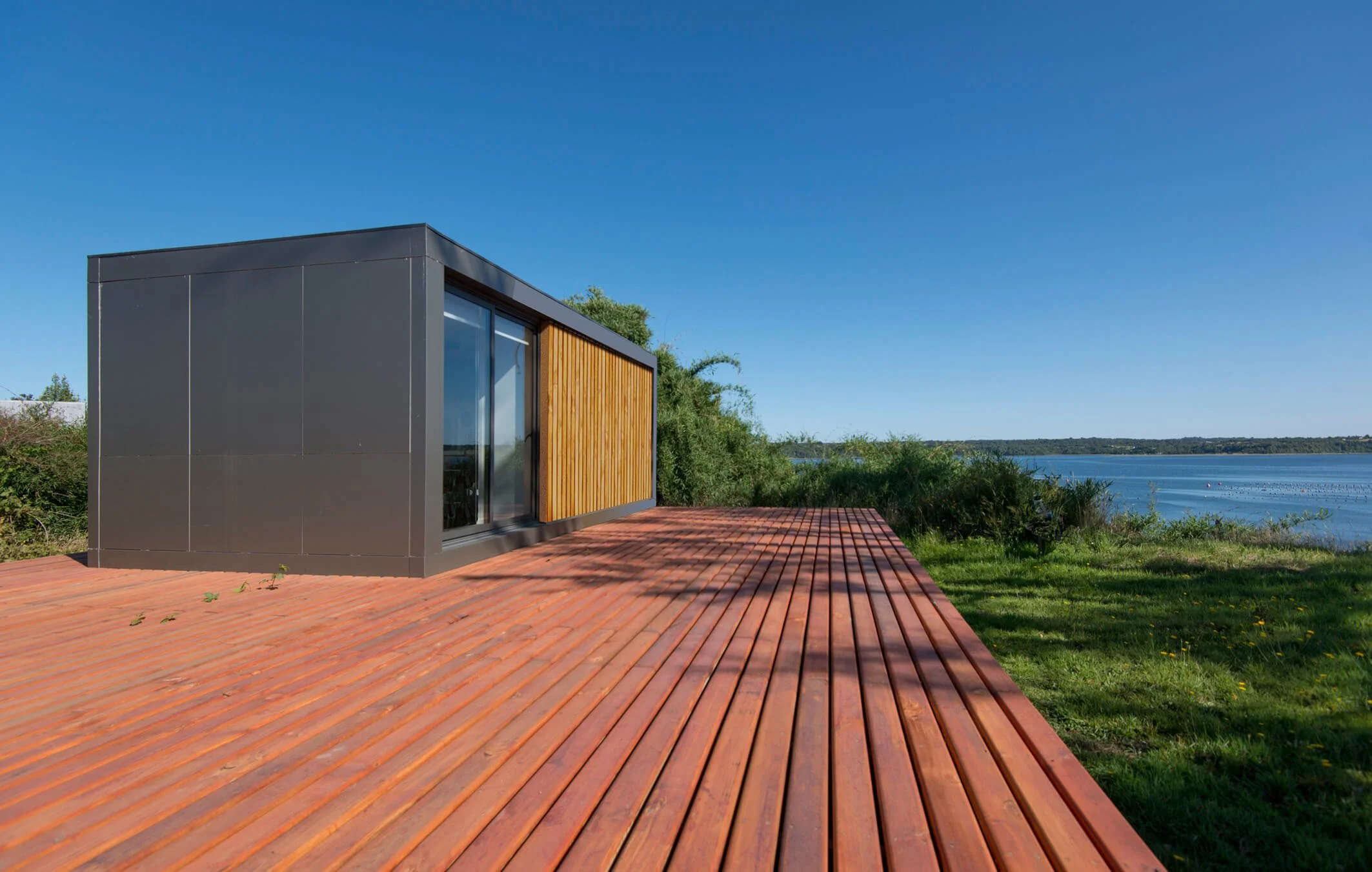 Casa moderna de diseño compacto y paredes negras, con puertas de cristal y detalles de madera, ubicada junto a un paisaje con lago y vegetación, en un día soleado y despejado.