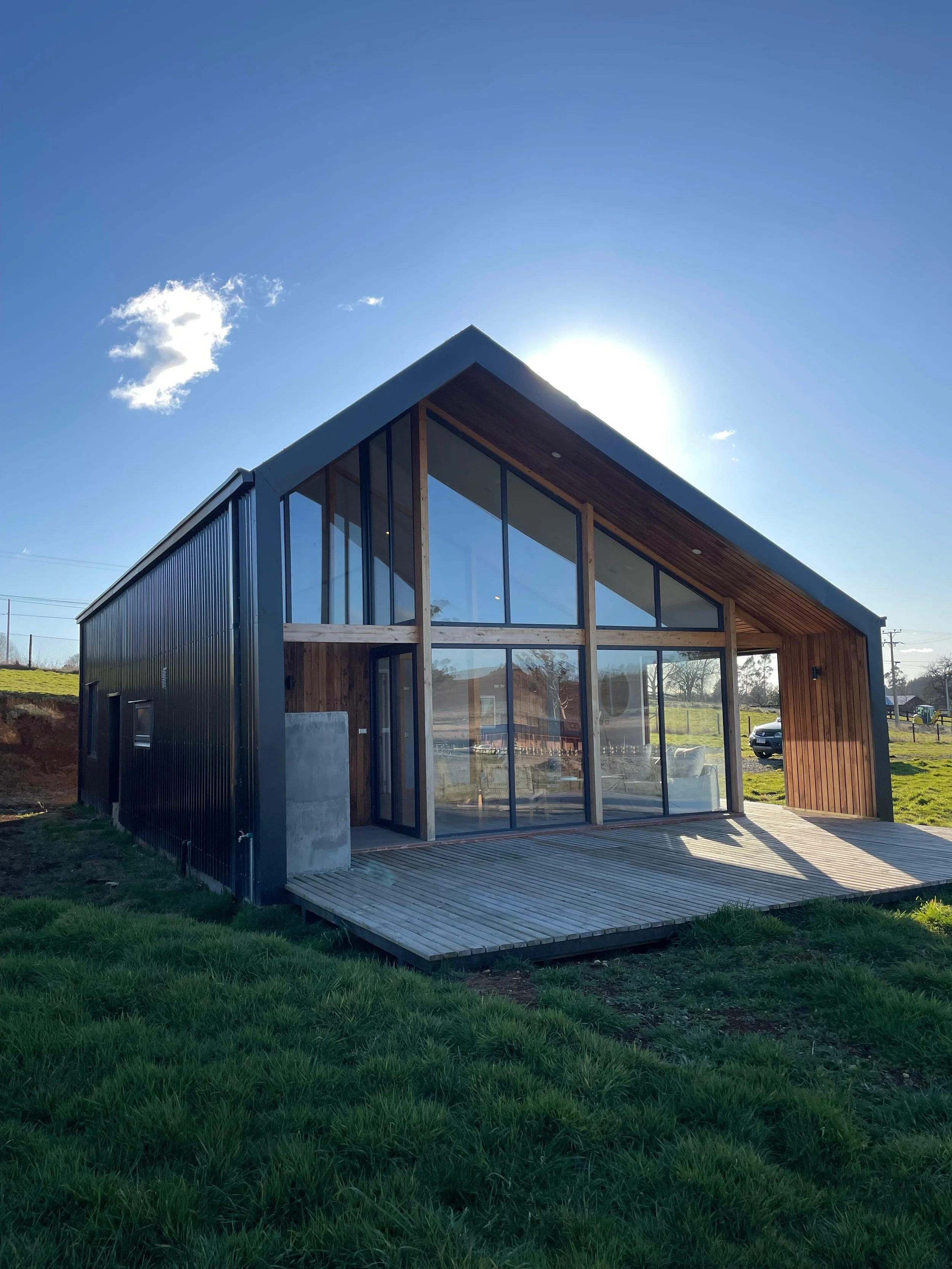 Casa moderna de estructura de madera y metal, con paredes de cristales grandes y un deck de madera, en un campo verde con cielo despejado.