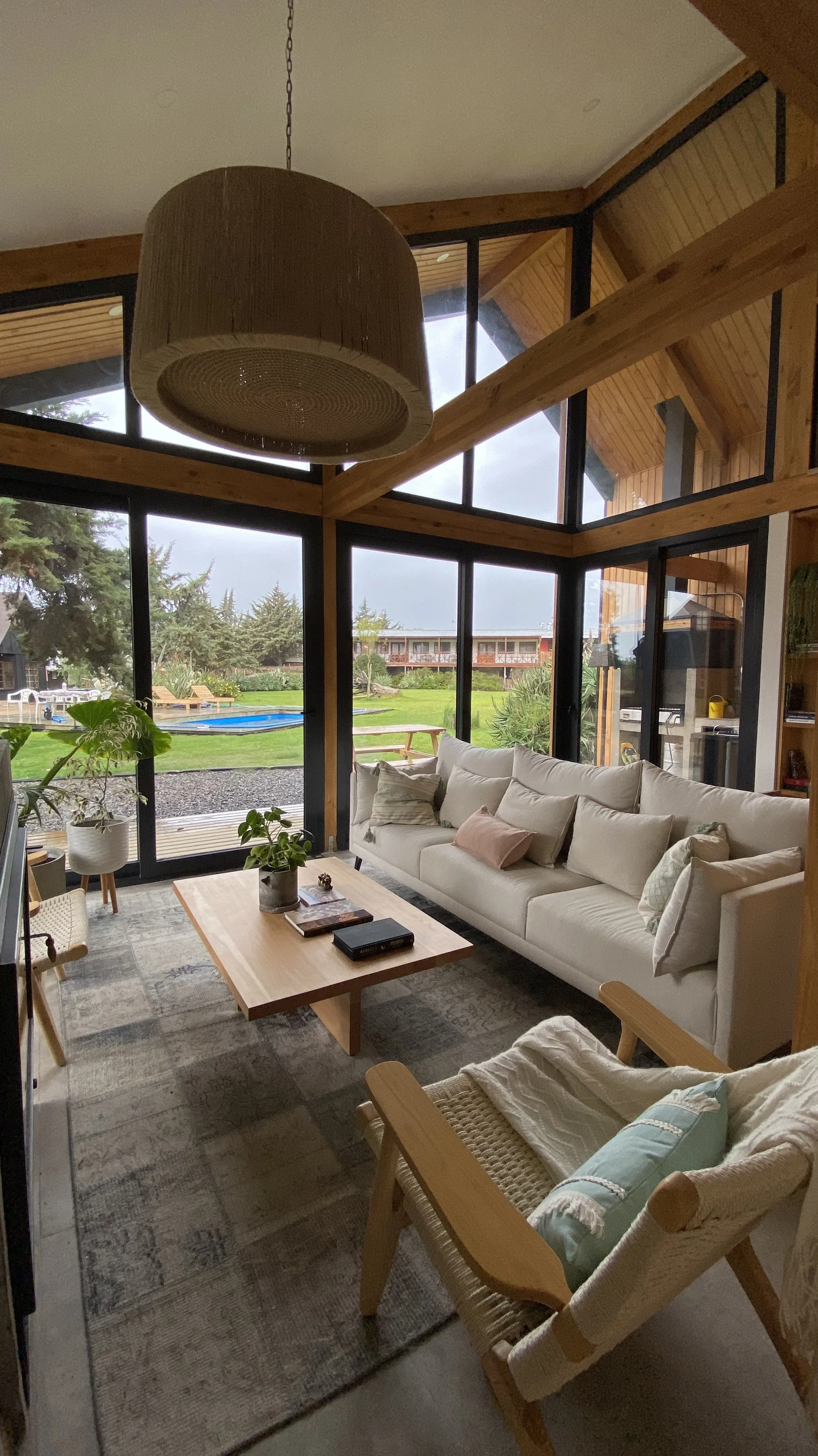 Sala de estar con grandes ventanas de vidrio, sofá blanco, mesa de madera, y vista hacia un jardín con piscina y césped.