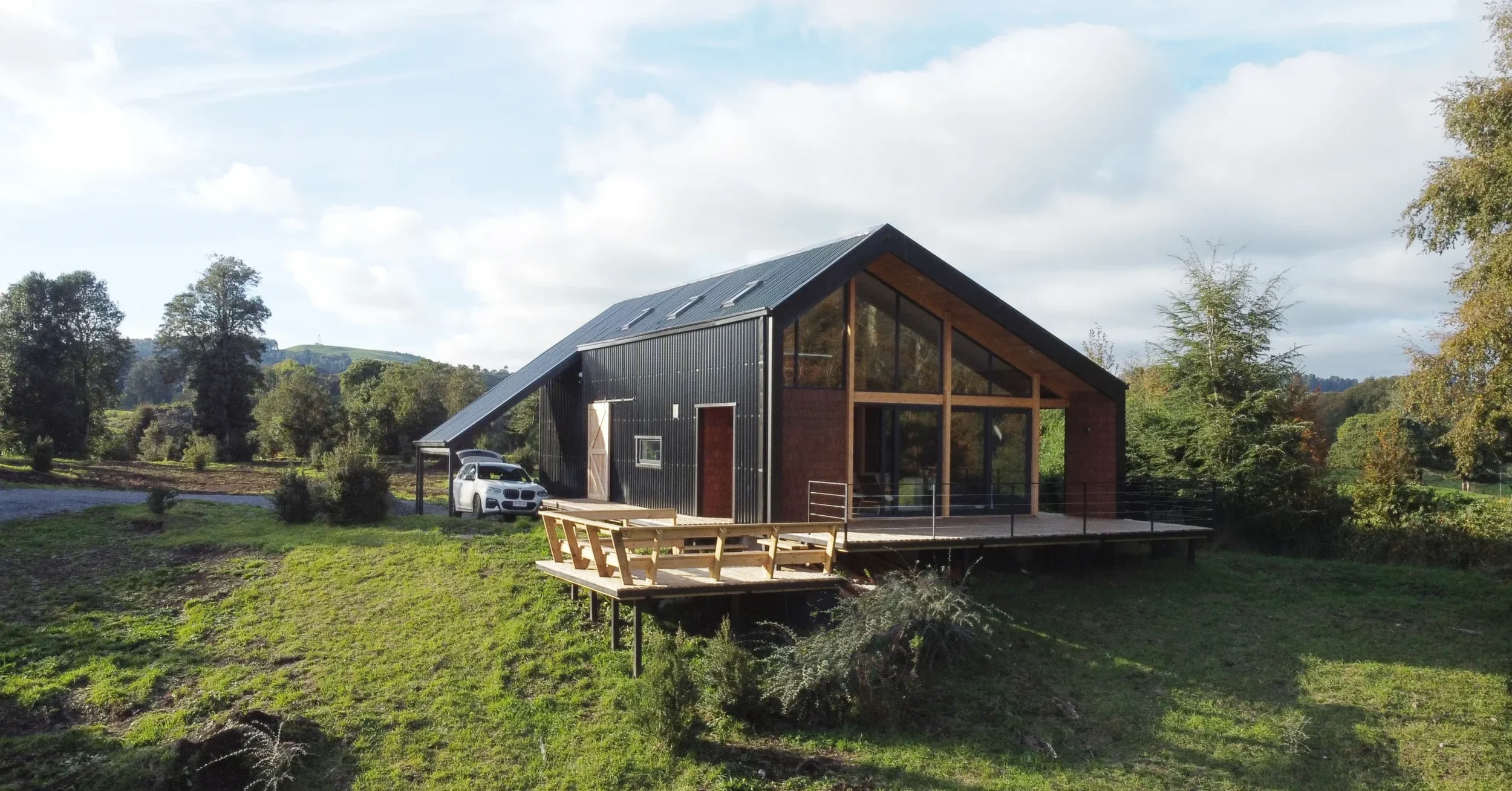 Una casa moderna en un entorno rural con árboles y prados verdes, con piezas de madera en el exterior y amplio balcón.