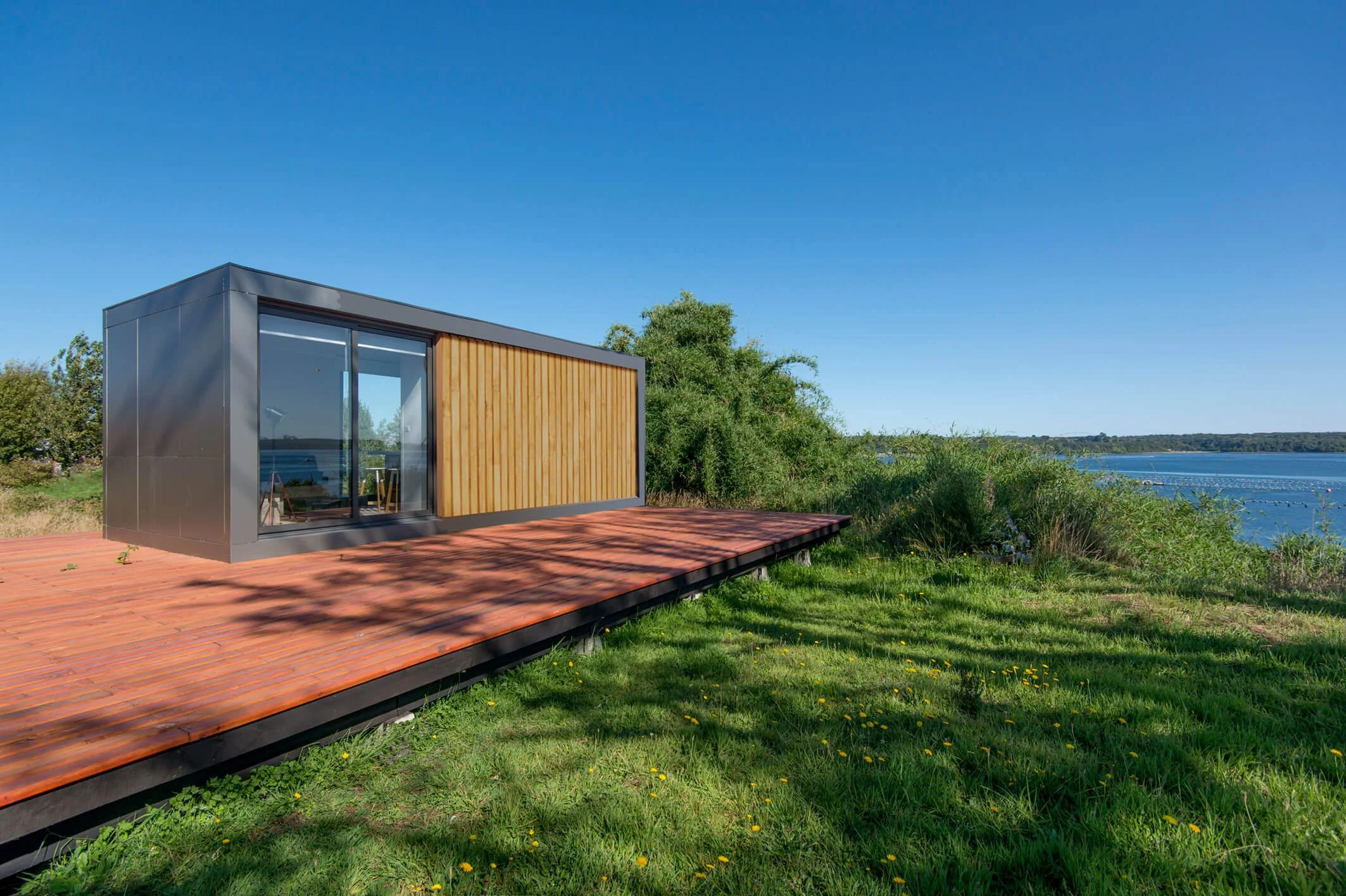 Casa moderna con paredes de madera y metal, ubicada junto a un lago, con un muelle de madera y césped verde en un día soleado.