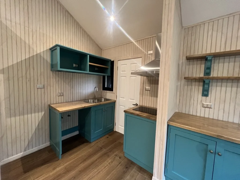 Cocina pequeña con gabinetes azules y encimera de madera, paredes de madera con rayas blancas y beige, estufa eléctrica y estantes de madera.