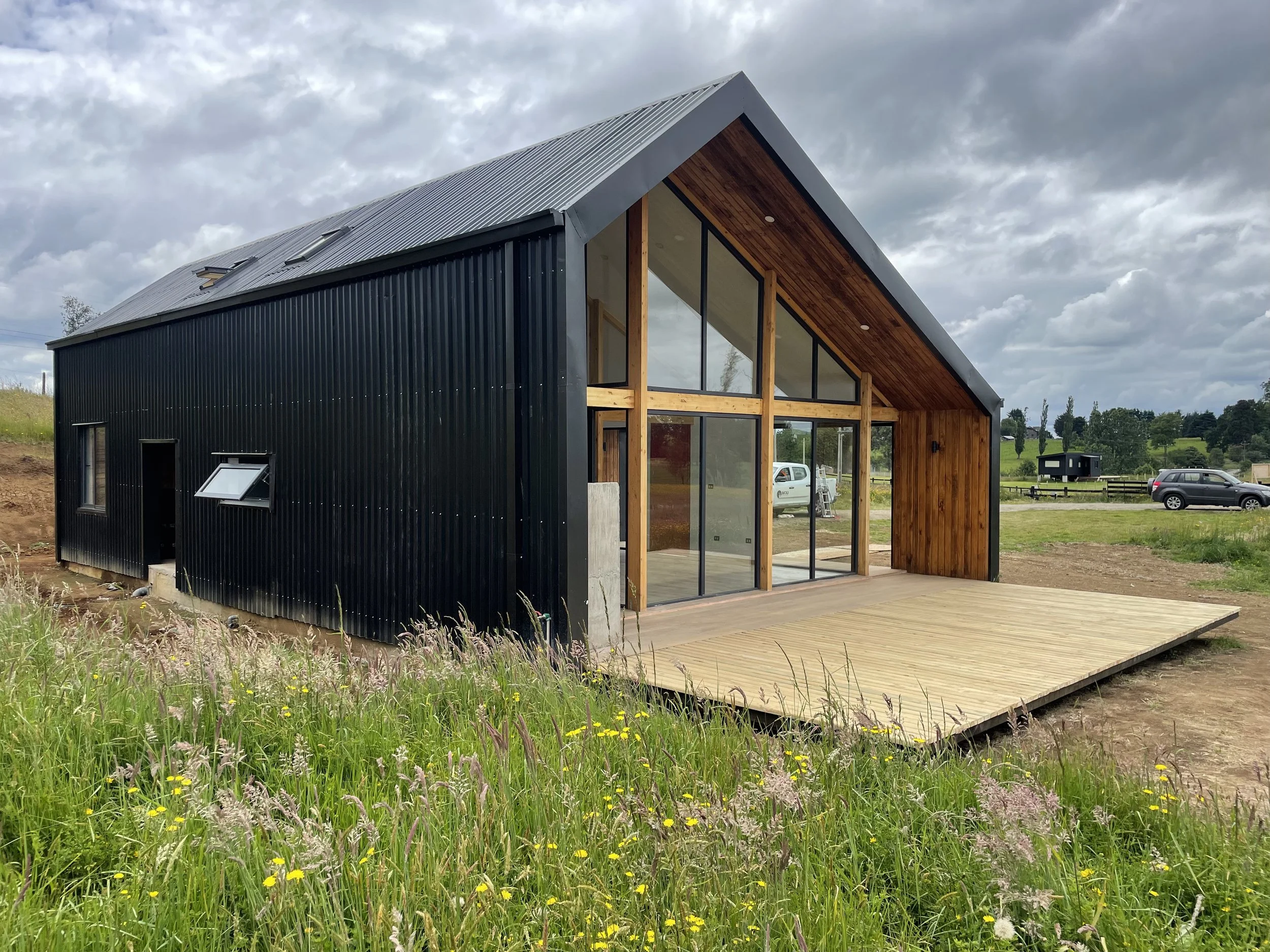 Casa moderna con estructura de madera y fachada de metal negra, con amplio deck de madera y grandes ventanas en un entorno rural con césped y caminos