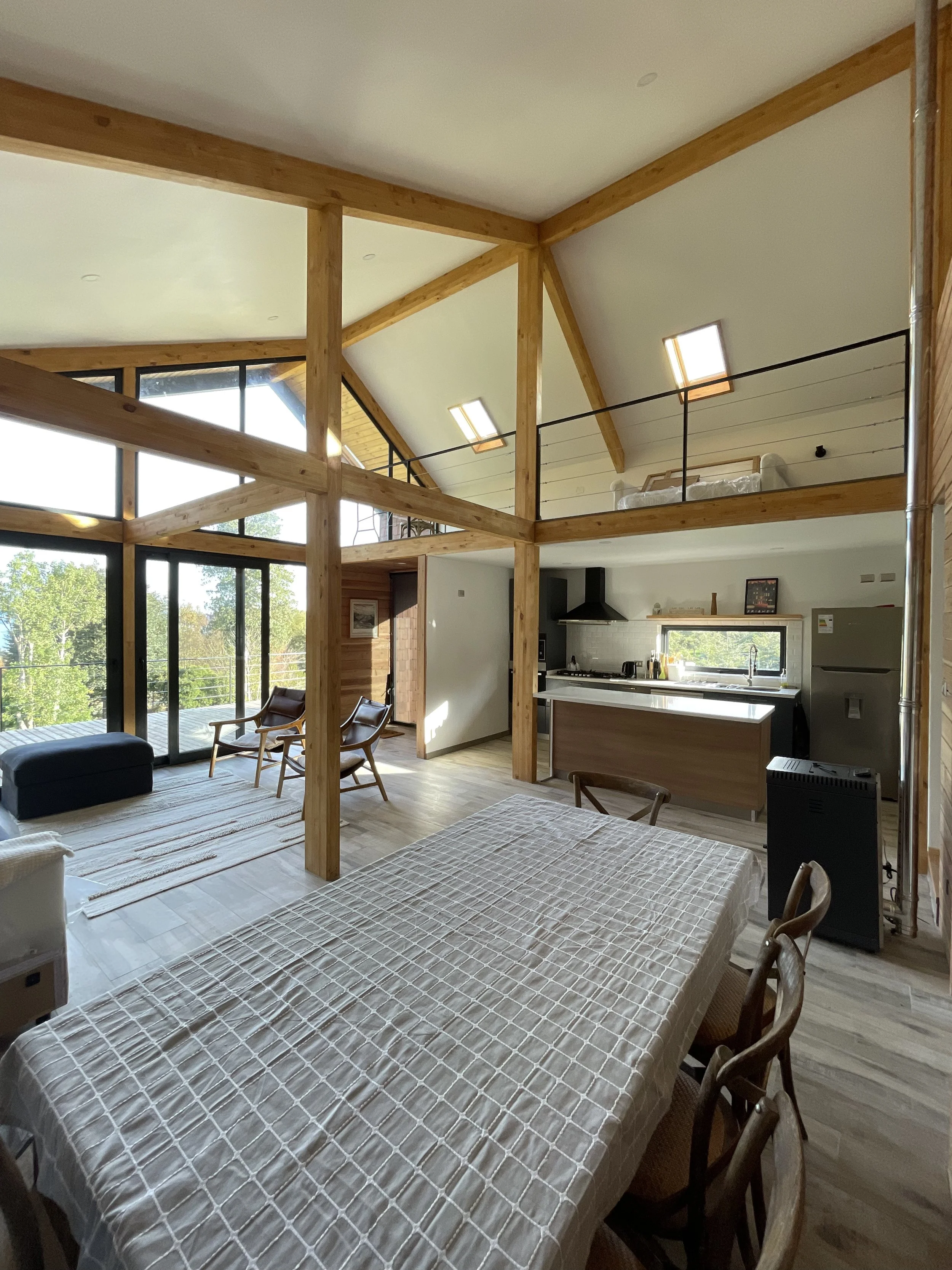 Interior de una casa moderna de techo alto con estructura de madera, sala con ventanas grandes, comedor y cocina abierta, muebles de madera y comedor con mantelería a cuadros.